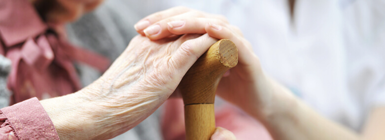 Elderly lady holding a wooden walking stick whilst a care home worker has a hand on top of her's