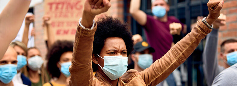 African-American-woman-wearing-protective-face-mask-while-protesting-with-arms-raised-on-city-streets