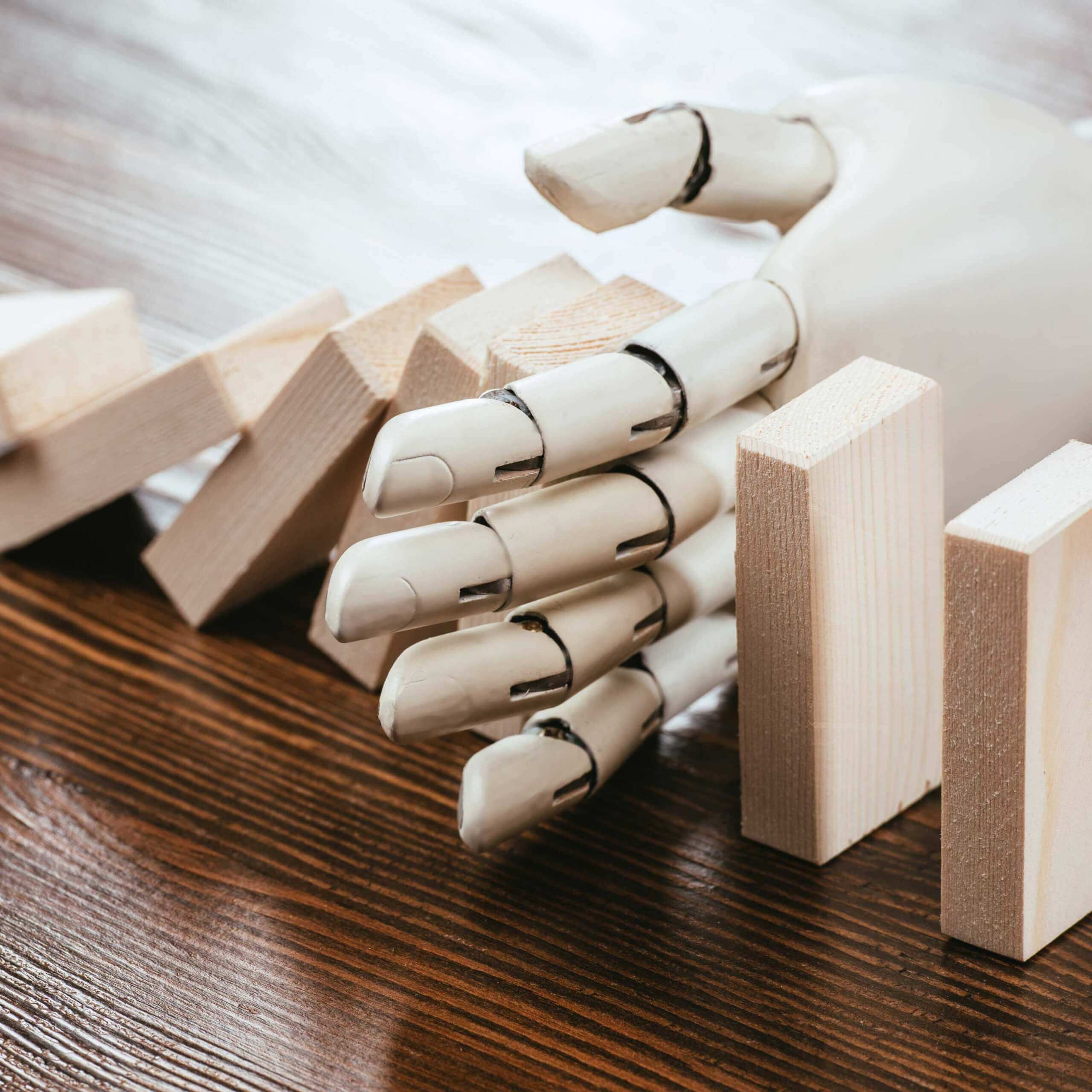 An image of a set of wooden dominoes, and what appears to be a robot hand blocking it. A visual metaphor for the topic of this post AI and recruitment: An invaluable tool, or source of discrimination.