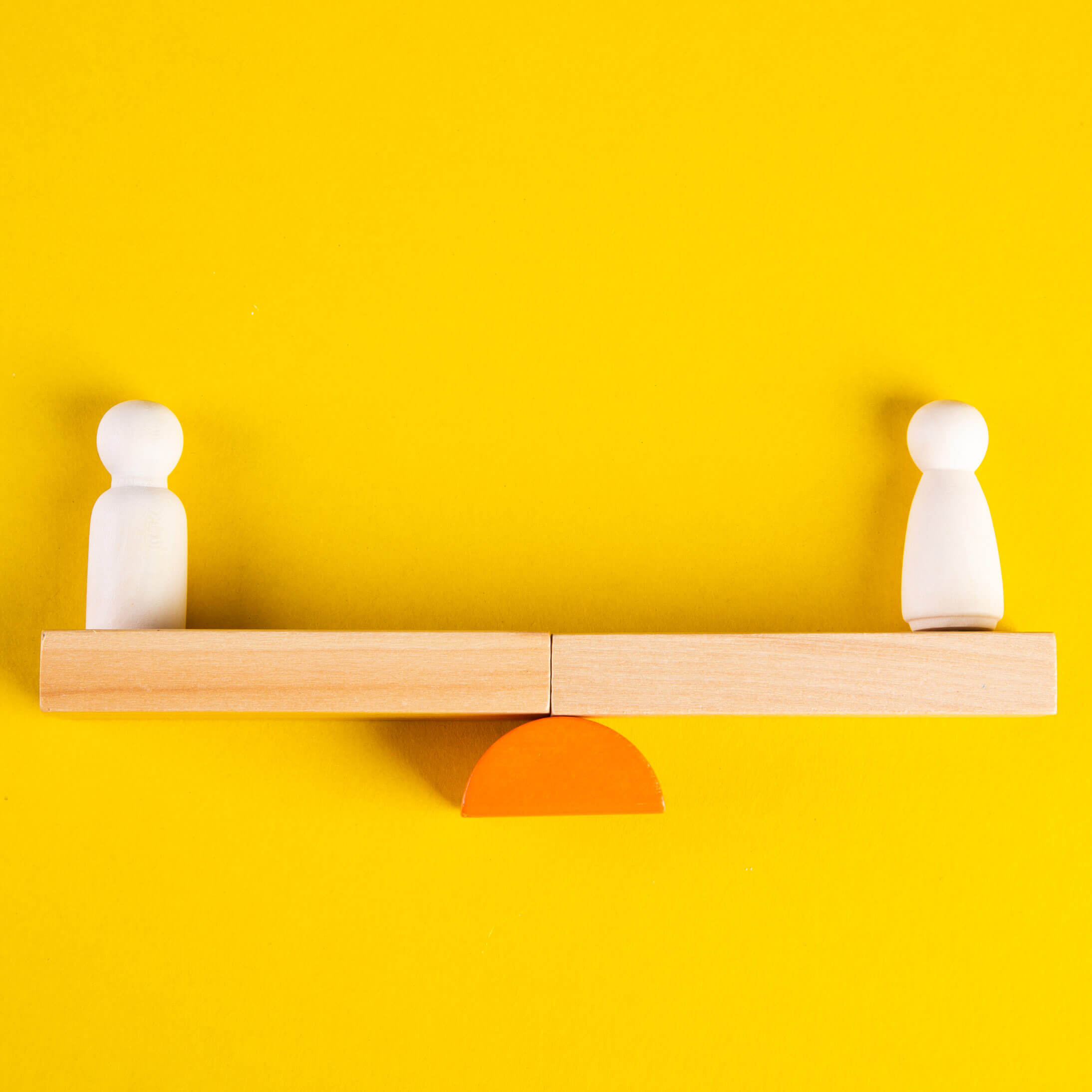 An image of two wooden figures on a wooden seesaw, placed flat on a yellow background. A visual metaphor for the topic of this article: The Worker Protection (Amendment of Equality Act 2010) Bill 2023.